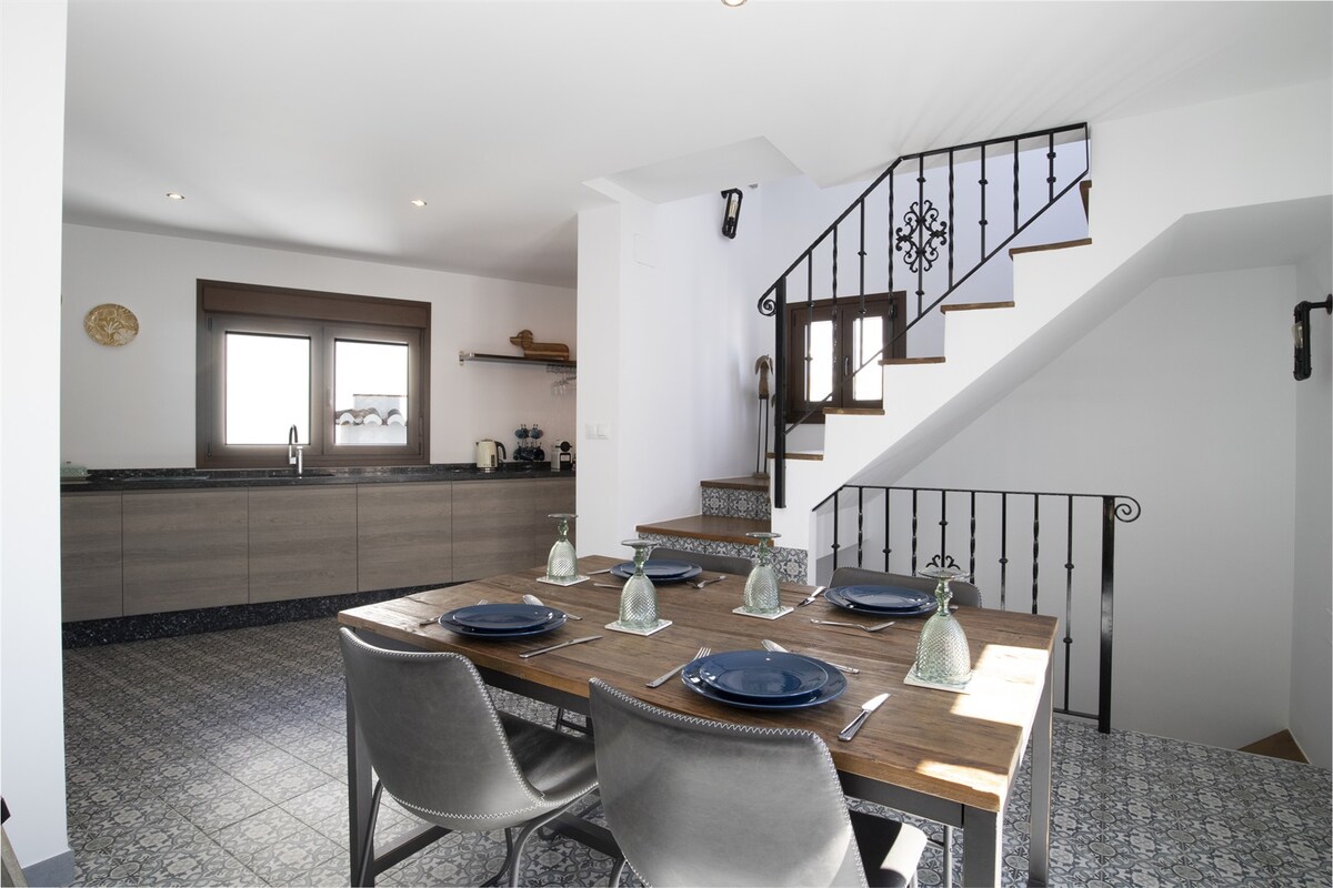 A modern dining area is positioned beside a well-equipped kitchen, featuring wooden furniture and tasteful table settings. Light-colored walls and tiled flooring enhance the space, while a staircase with a wrought-iron railing leads to the upper level, creating a sense of depth.