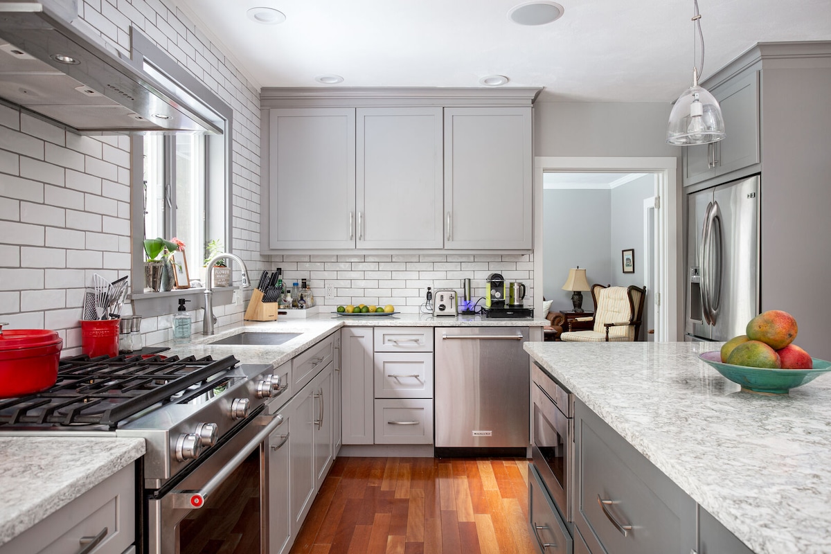 A modern kitchen features grey cabinetry and a marble countertop, complemented by glossy subway tile backsplash. Stainless steel appliances, including a gas stove and dishwasher, are positioned along the walls. A bowl of fresh fruit adds a pop of color on the counter.