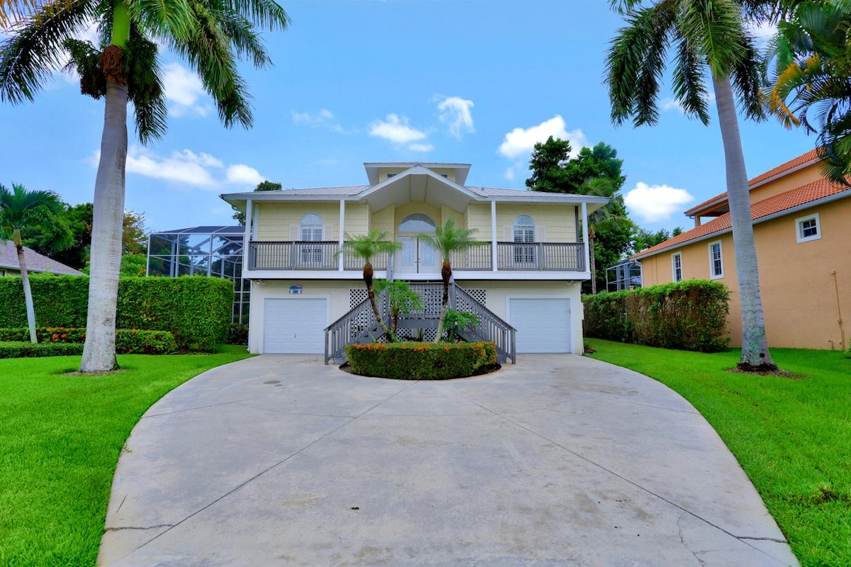 A spacious home is positioned on a wide driveway with a circular layout. Lush green grass surrounds the property, complemented by palm trees. A covered entrance is framed by elegant arches, and garages are located beneath the house, providing ample parking space.