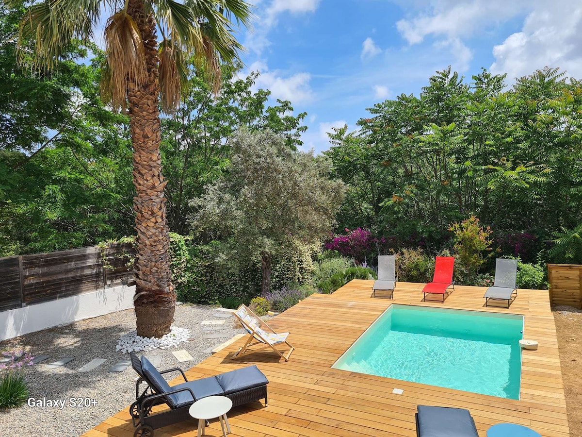 A private pool is surrounded by a wooden deck, complemented by lounge chairs in contrasting colors. Lush greenery and blooming plants create a vibrant backdrop. A tall palm tree provides shade, contributing to a serene outdoor space ideal for relaxation.