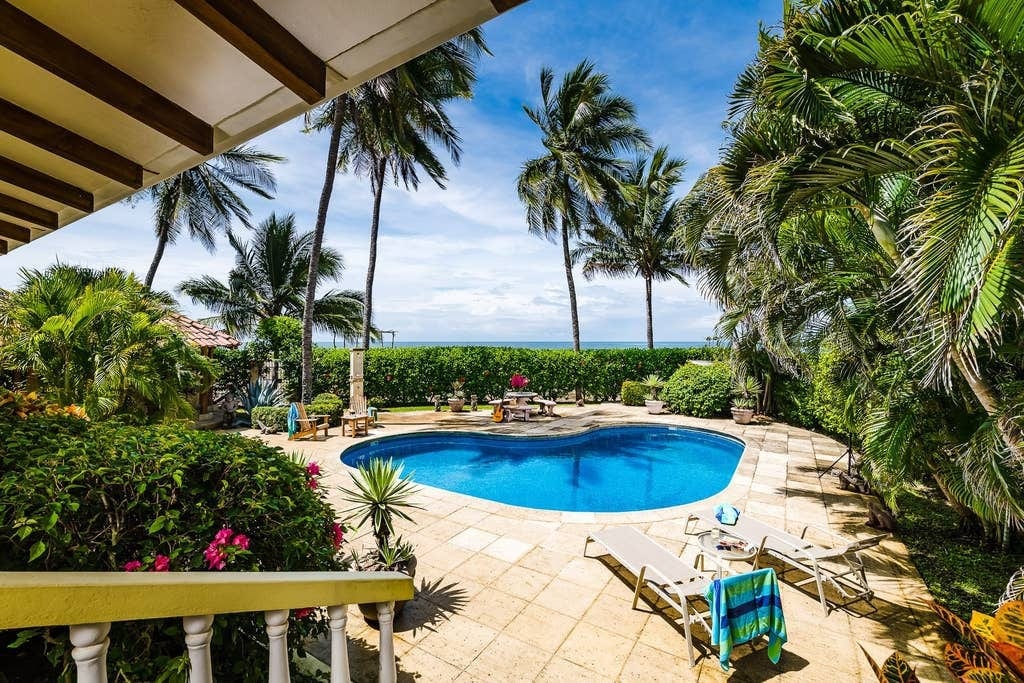 A private pool is surrounded by lush greenery and palm trees, creating a tropical oasis. Sun loungers are positioned along the poolside, with a colorful towel draped over one. The blue water reflects the clear sky, adding a serene atmosphere to the outdoor space.