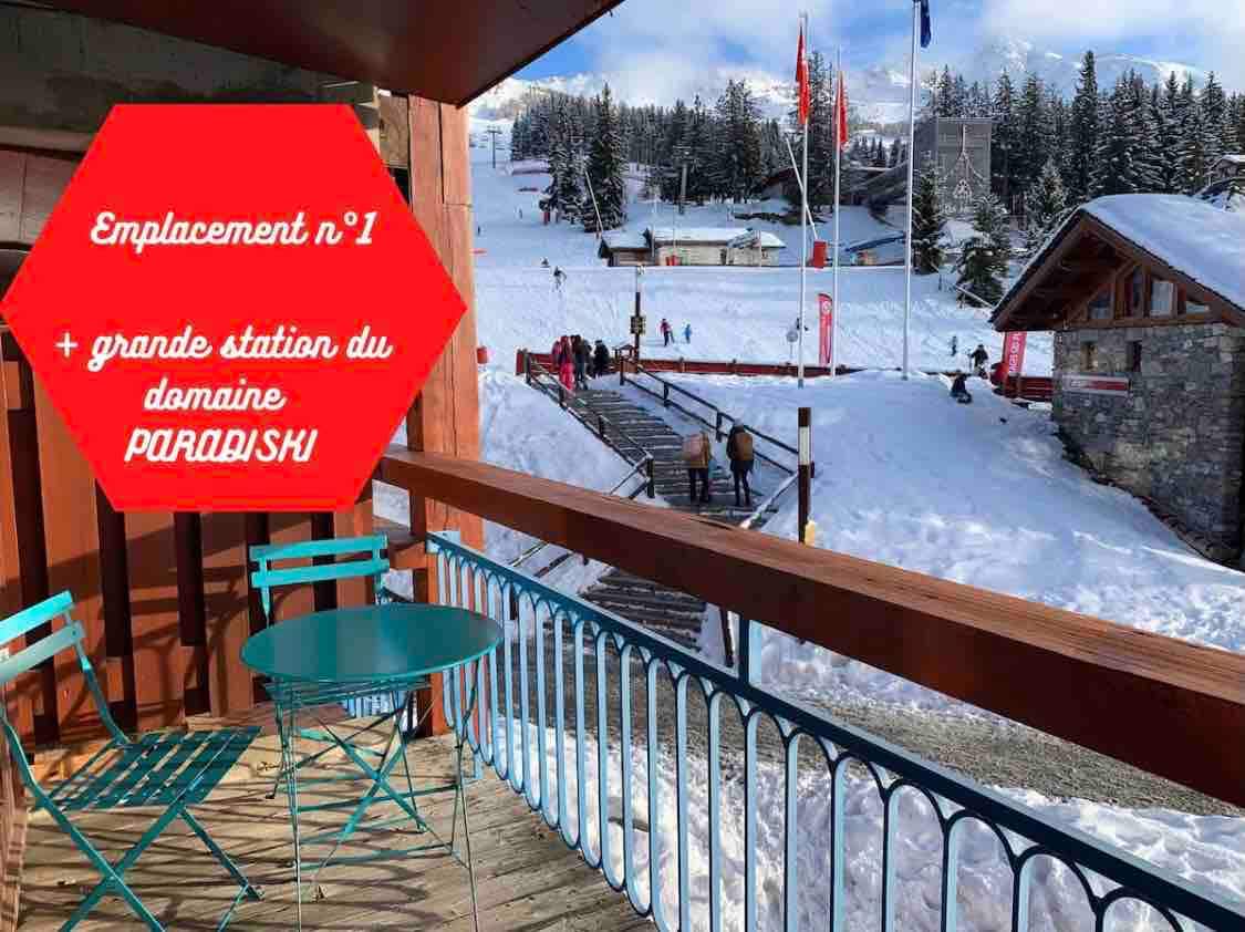 A balcony view showcases a snow-covered landscape and ski slopes. The scene is complemented by a cozy outdoor seating area, with two chairs and a small table. Skiers can be seen in the distance, enjoying the winter activities at the Paradiski resort.