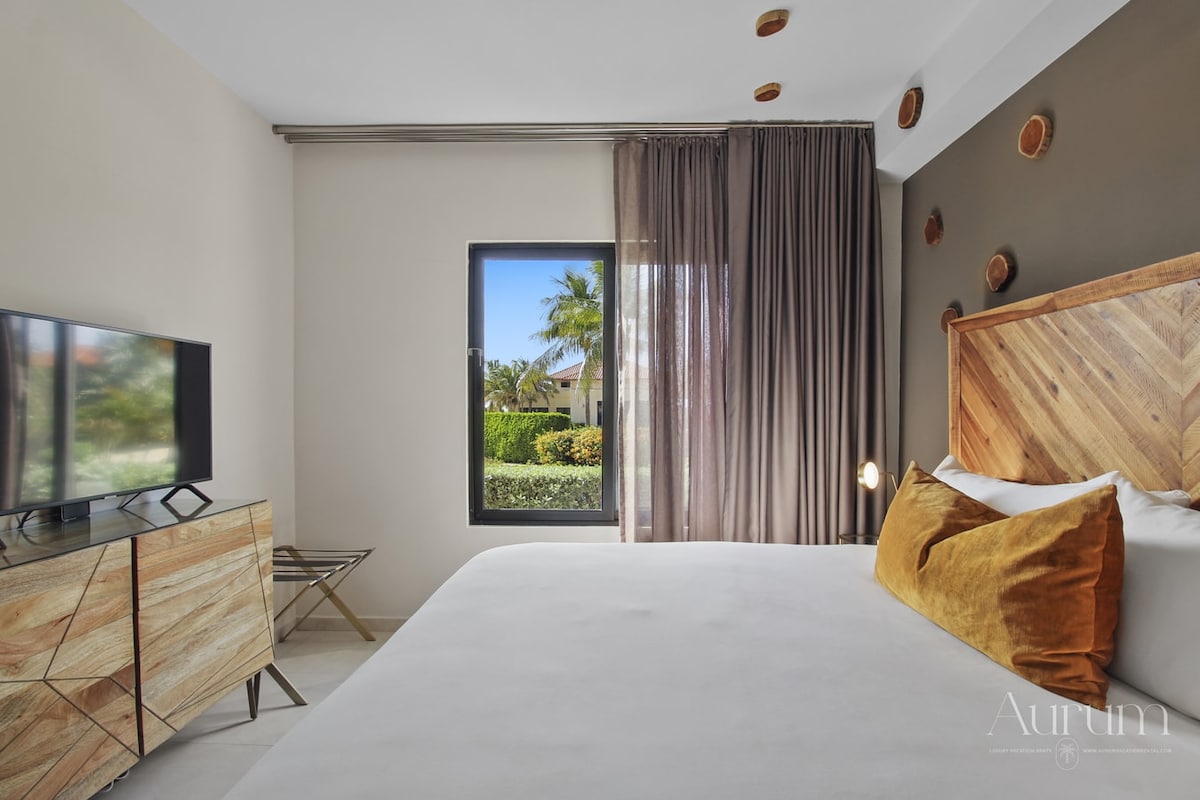 A spacious bedroom features a large bed topped with a soft white duvet and accentuated by decorative pillows. A modern wooden headboard complements the earthy tones. Natural light filters through the sheer curtains, illuminating the room, which also includes a flat-screen TV and a contemporary side table.