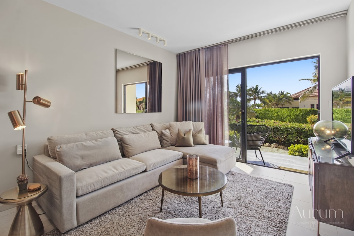 A comfortable living area is presented with a plush sectional sofa, accompanied by a round coffee table. Large glass doors allow natural light to fill the space, revealing views of lush greenery outside. A stylish mirror and decorative accents enhance the modern design.