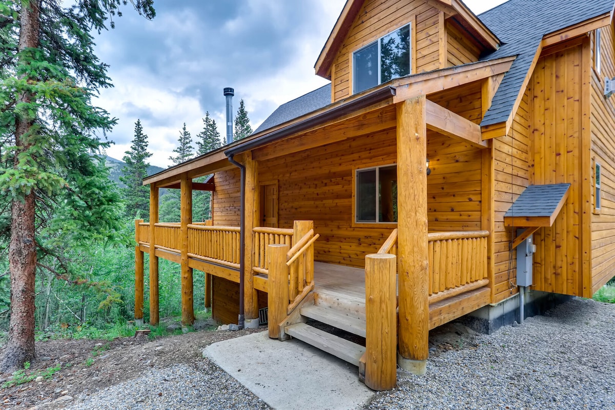 A spacious log home exterior is shown, featuring a large front porch with wooden railings and steps leading to a gravel pathway. The warm wood tones of the house contrast with the surrounding greenery of pine trees, creating a secluded atmosphere.