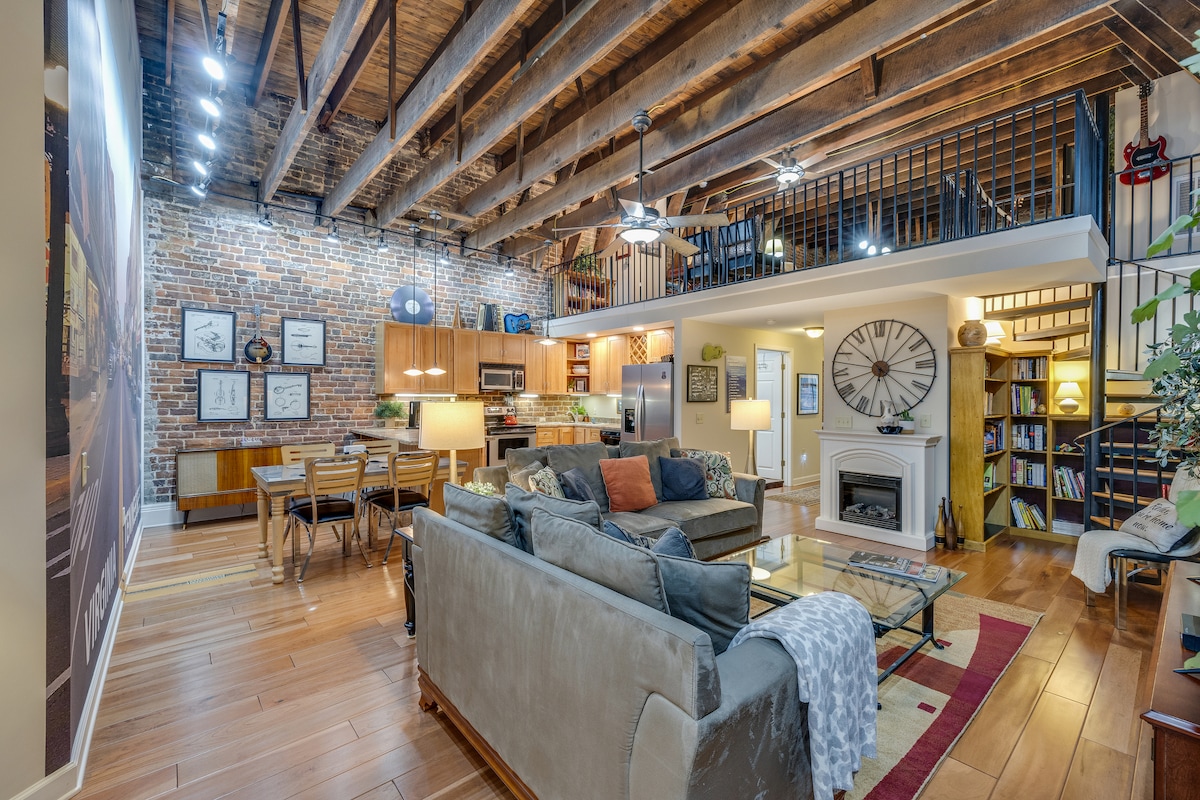 An open-concept living area showcases exposed wooden beams and a brick wall, enhancing the loft's historic character. A comfortable seating arrangement facing a glass coffee table offers a relaxed atmosphere. The gourmet kitchen features custom cabinetry, while a large clock adds visual interest above a cozy fireplace.