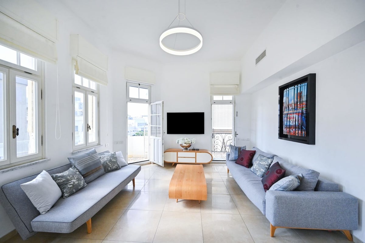 A bright living room features two modern sofas adorned with decorative pillows. A wooden coffee table sits centrally, while a flat screen TV is mounted on the wall. Large windows allow natural light to fill the space, and gray tiles enhance the contemporary feel.