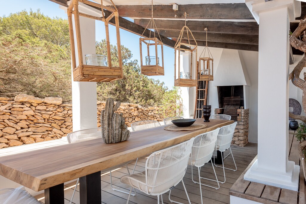 An outdoor dining area features a long wooden table surrounded by six white wire chairs. Lanterns are suspended overhead, and a stone wall provides a natural backdrop. A traditional fireplace is located to one side, enhancing the inviting outdoor space.