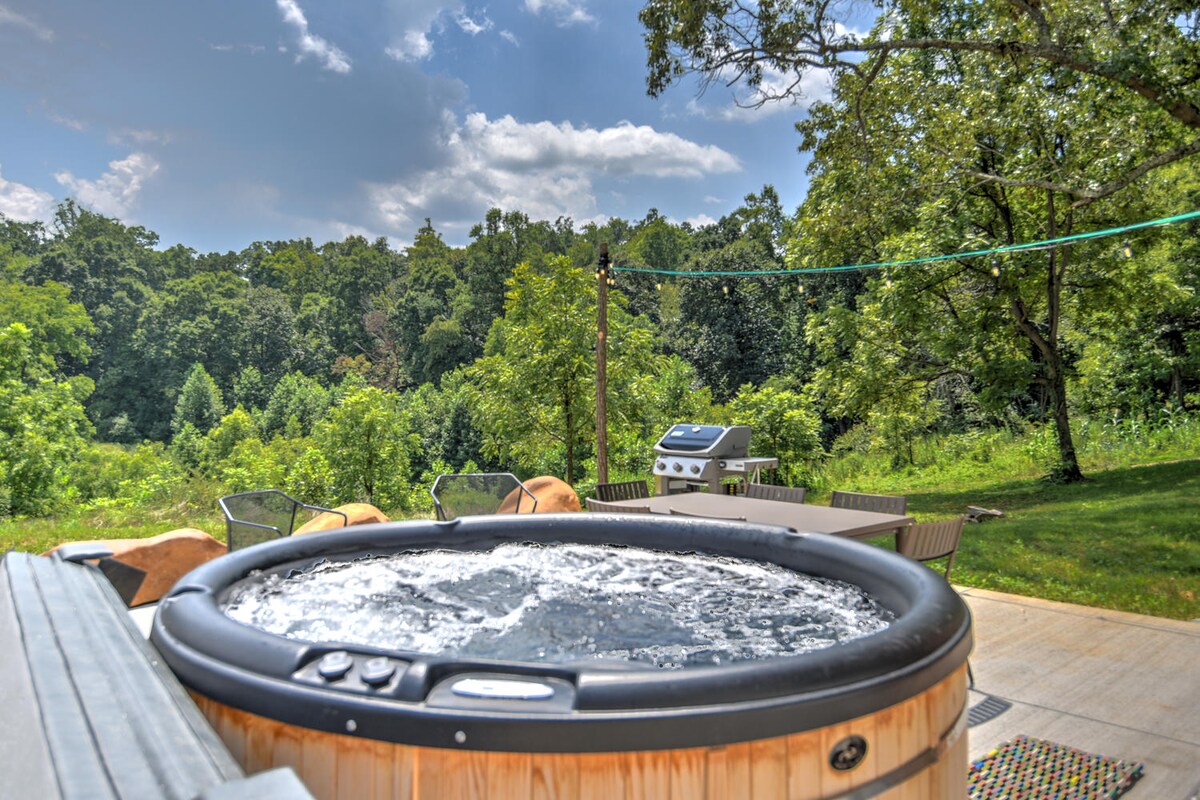 An outdoor hot tub is surrounded by lush greenery, providing a serene setting. A barbecue grill can be seen nearby, with several lounge chairs positioned to enjoy the natural views. Clear blue sky and soft clouds contribute to the tranquil atmosphere.
