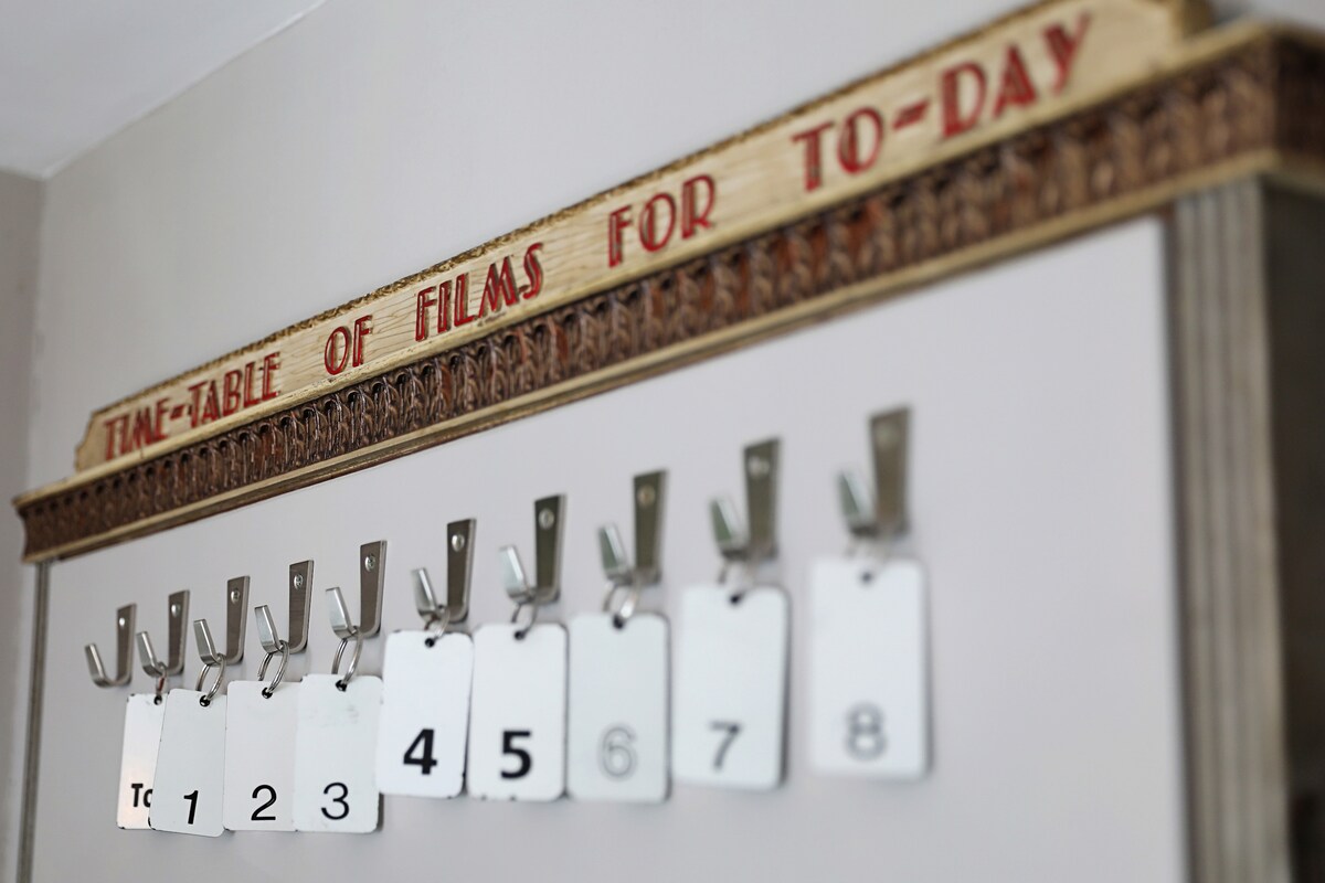 A decorative board is mounted on the wall, featuring the title 'TIME-TABLE OF FILMS FOR TO-DAY.' Below, several metal hooks are displayed, holding blank tags numbered from one to eight, indicating a film schedule.