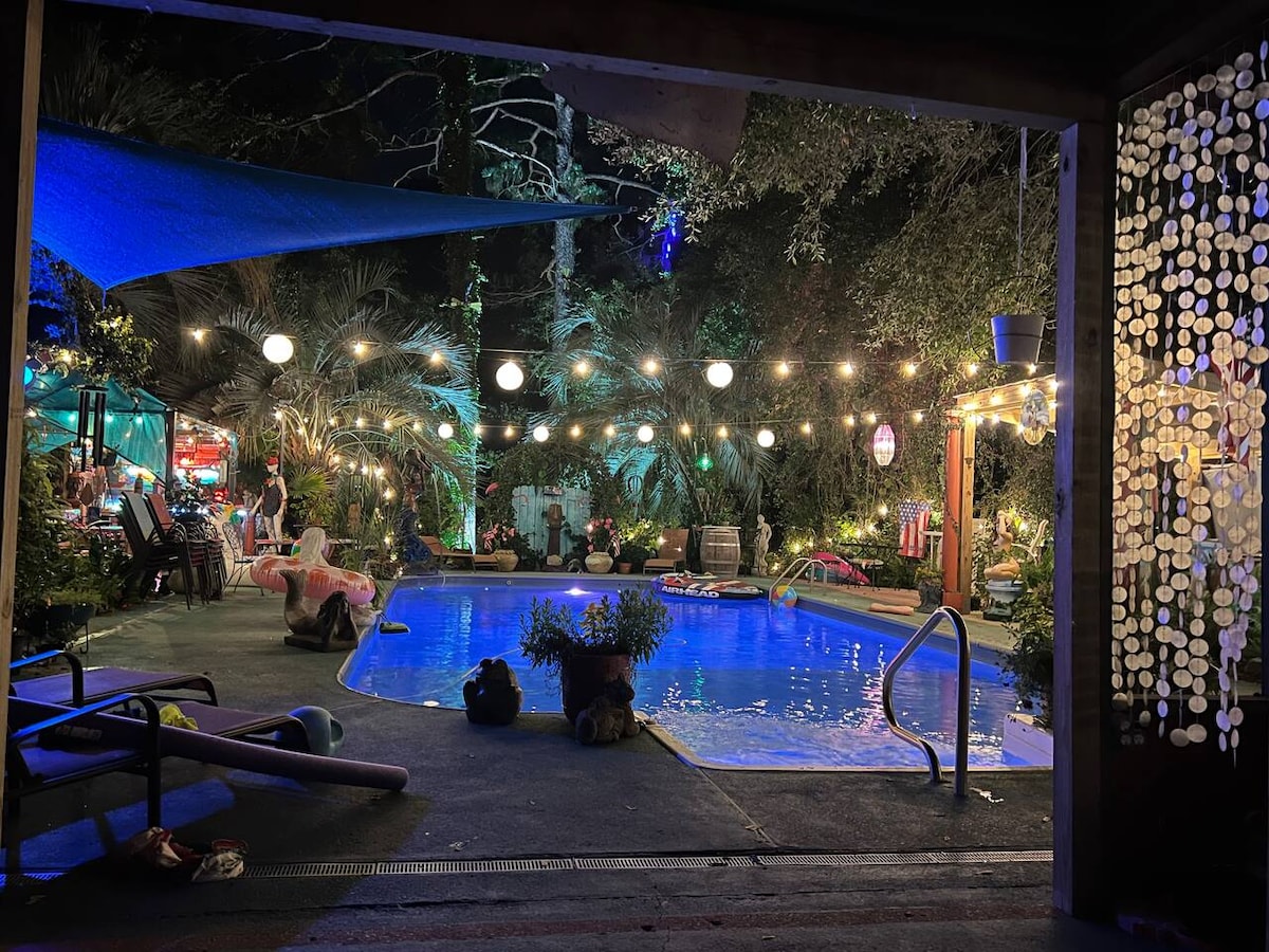 An inviting pool area features a sparkling blue water pool surrounded by lush greenery and illuminated by soft, ambient lights. Lounge chairs are positioned nearby, and decorative plants add color. A shaded area is visible, along with playful pool floats, creating a relaxed atmosphere.