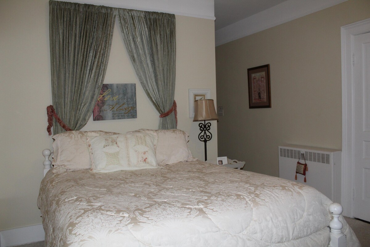 A spacious bed, adorned with a light-colored quilt, is framed by elegant drapes that create a soft ambiance. A decorative piece hangs above the bed, and a small lamp stands beside it, adding subtle lighting to the cozy atmosphere.
