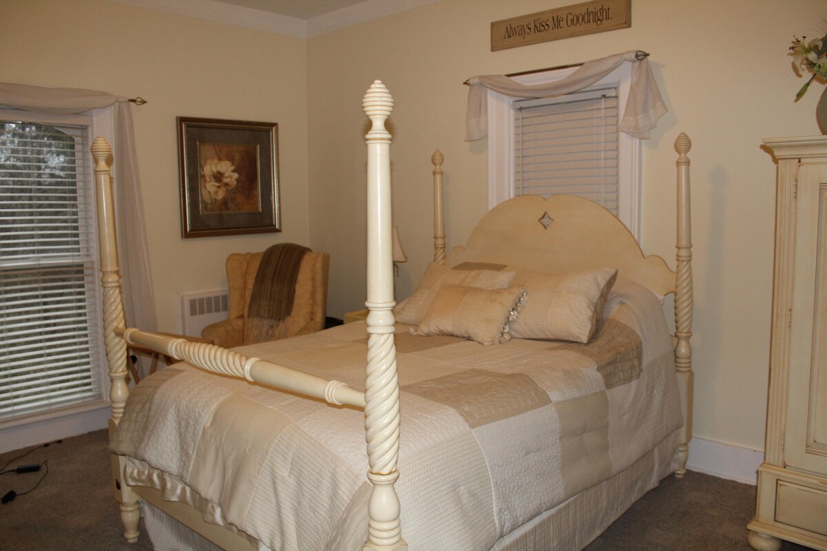 A bright room features a four-poster queen bed adorned with neutral-colored bedding and decorative pillows. A comfortable armchair is positioned nearby, and natural light filters through the window, enhancing the soft yellow walls.
