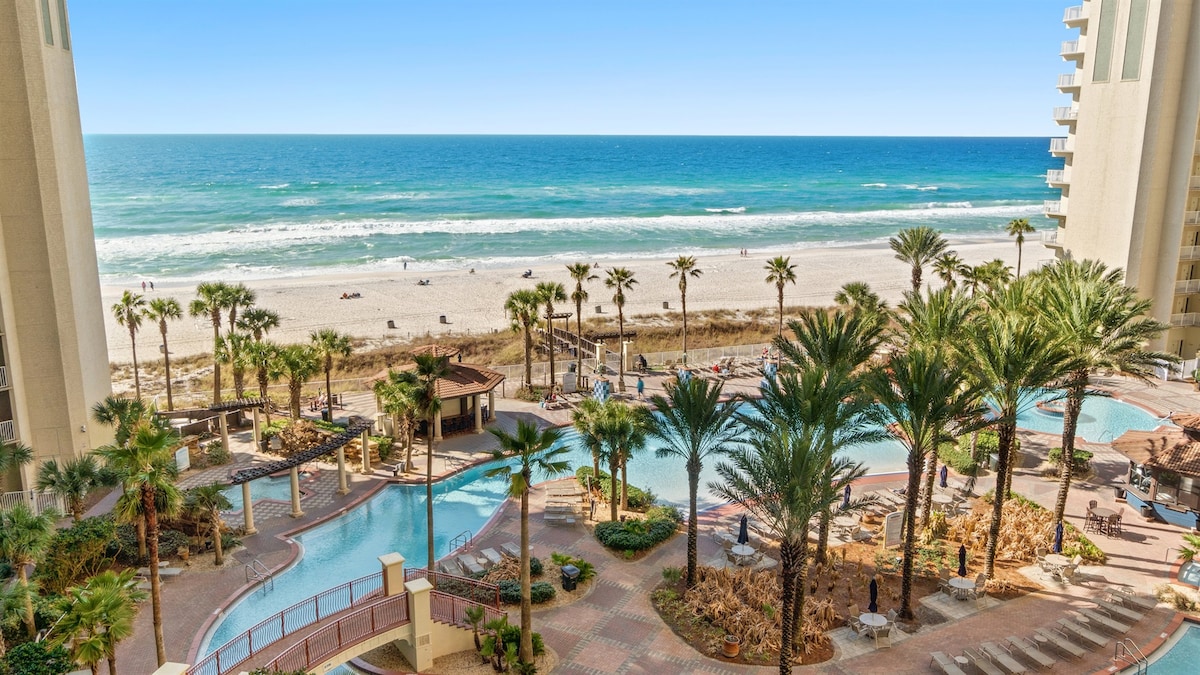 A view from the balcony highlights the expansive beach and ocean beyond. Lush palm trees frame the resort's lagoon pool area, which is surrounded by lounge chairs and a gazebo. The water glistens under the clear blue sky, providing a tranquil backdrop.
