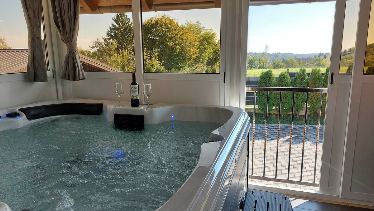 A hot tub is positioned beside large windows that offer views of a verdant landscape. Glasses and a bottle are set on the edge of the tub, enhancing the setting for relaxation. Natural light fills the space, creating a serene atmosphere.