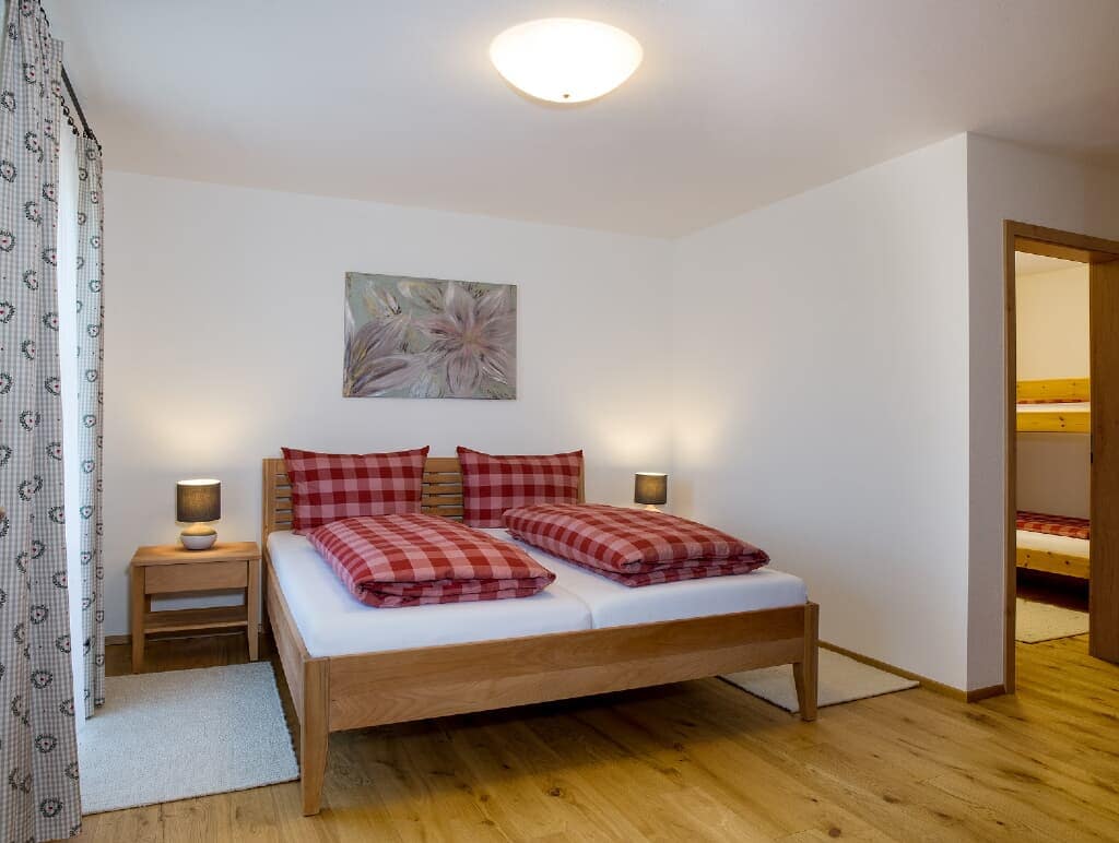 A cozy bedroom features a spacious bed with a checkered red and white duvet. Two bedside lamps provide soft lighting, while a piece of floral wall art adds a touch of decor. Light wooden flooring complements the gentle color palette.