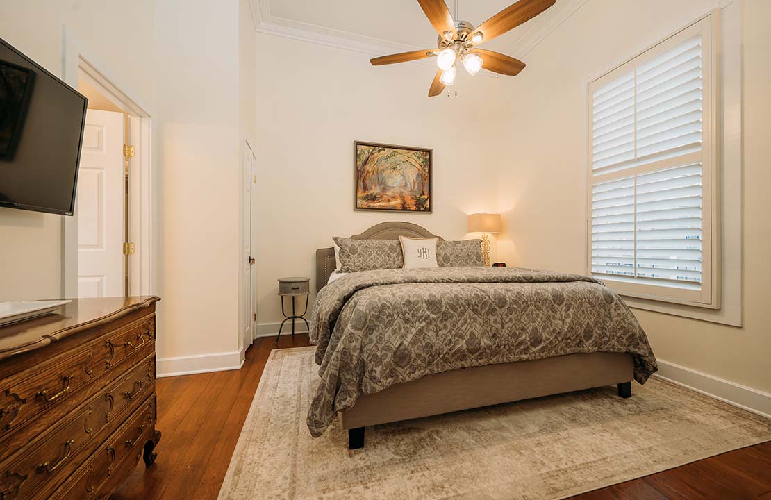 A spacious bedroom features a large bed dressed in neutral-toned bedding. Natural light enters through a well-placed window with blinds. A ceiling fan and a soft area rug add comfort, while a wooden dresser and framed artwork provide subtle accents.