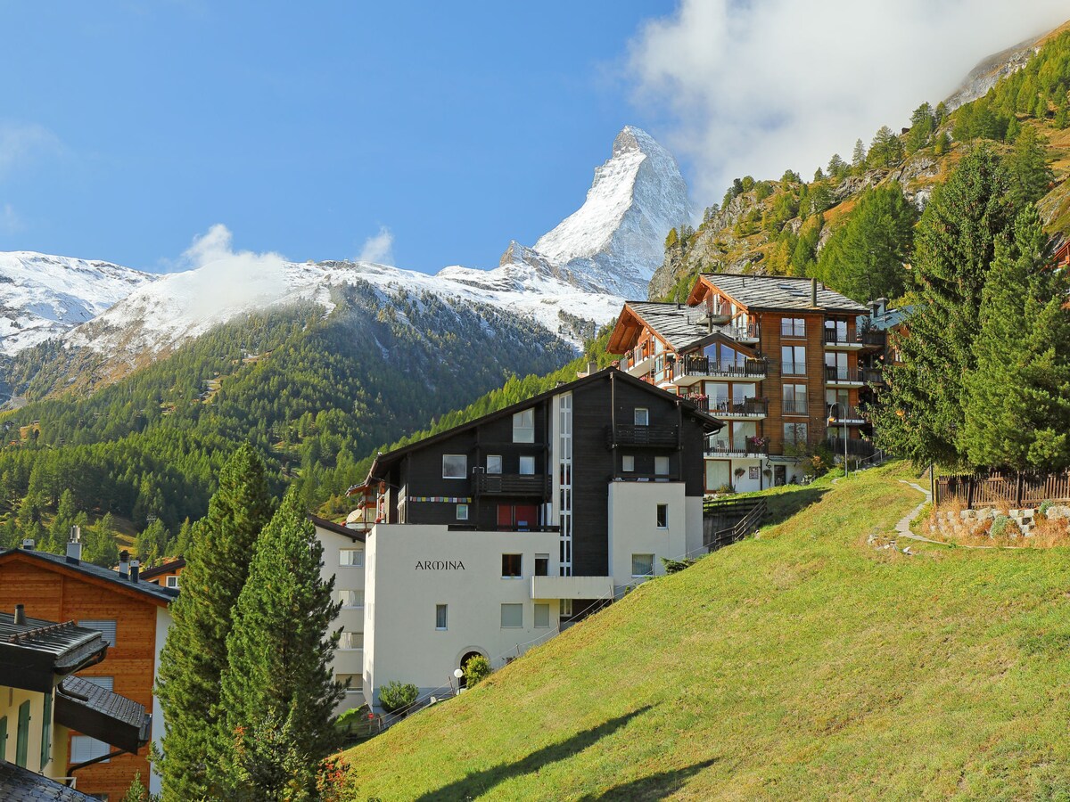 The chalet-style apartment house 'Armina' is nestled on a hillside, surrounded by lush greenery. The iconic Matterhorn peak rises majestically in the background, partially shrouded in clouds. Modern mountain architecture complements the natural landscape.