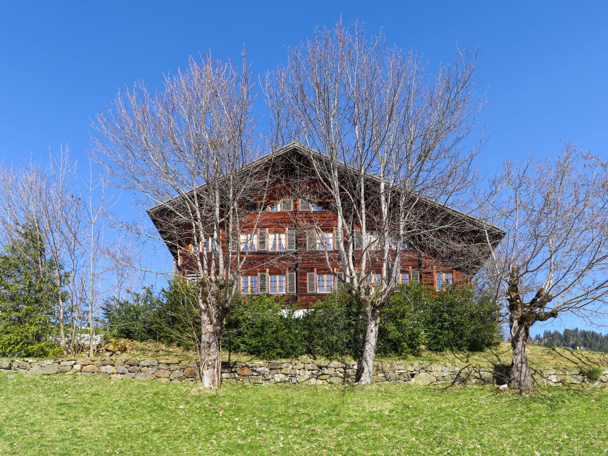 A three-story wooden chalet is surrounded by a grassy area and several bare trees. The building showcases a traditional design with multiple windows and a sloped roof, against a clear blue sky.