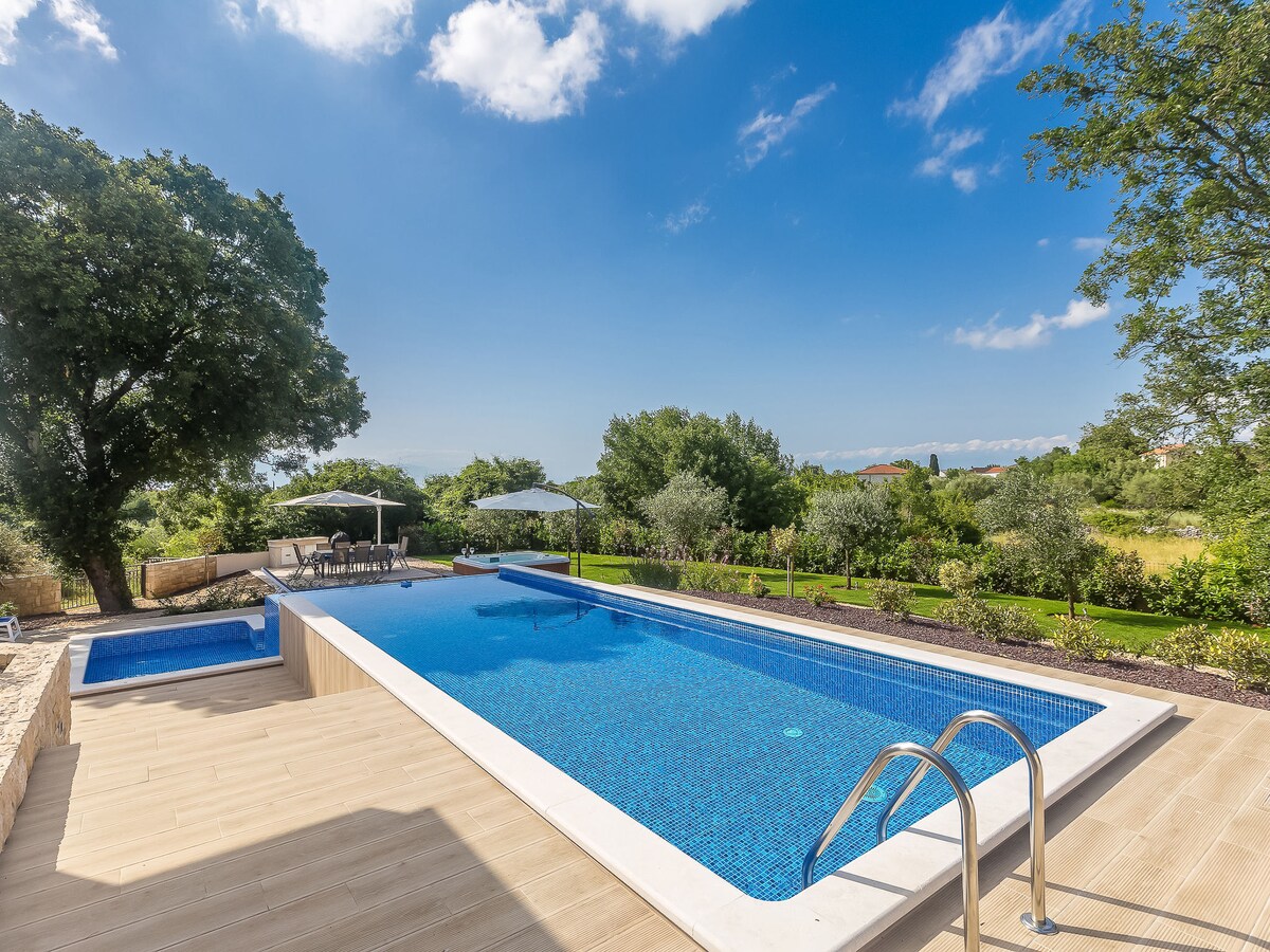 An angular swimming pool with a vibrant blue surface is surrounded by a wooden deck. Sun loungers and umbrellas provide comfortable seating options. Lush greenery, including trees and shrubs, frames the area, contributing to a serene outdoor atmosphere.