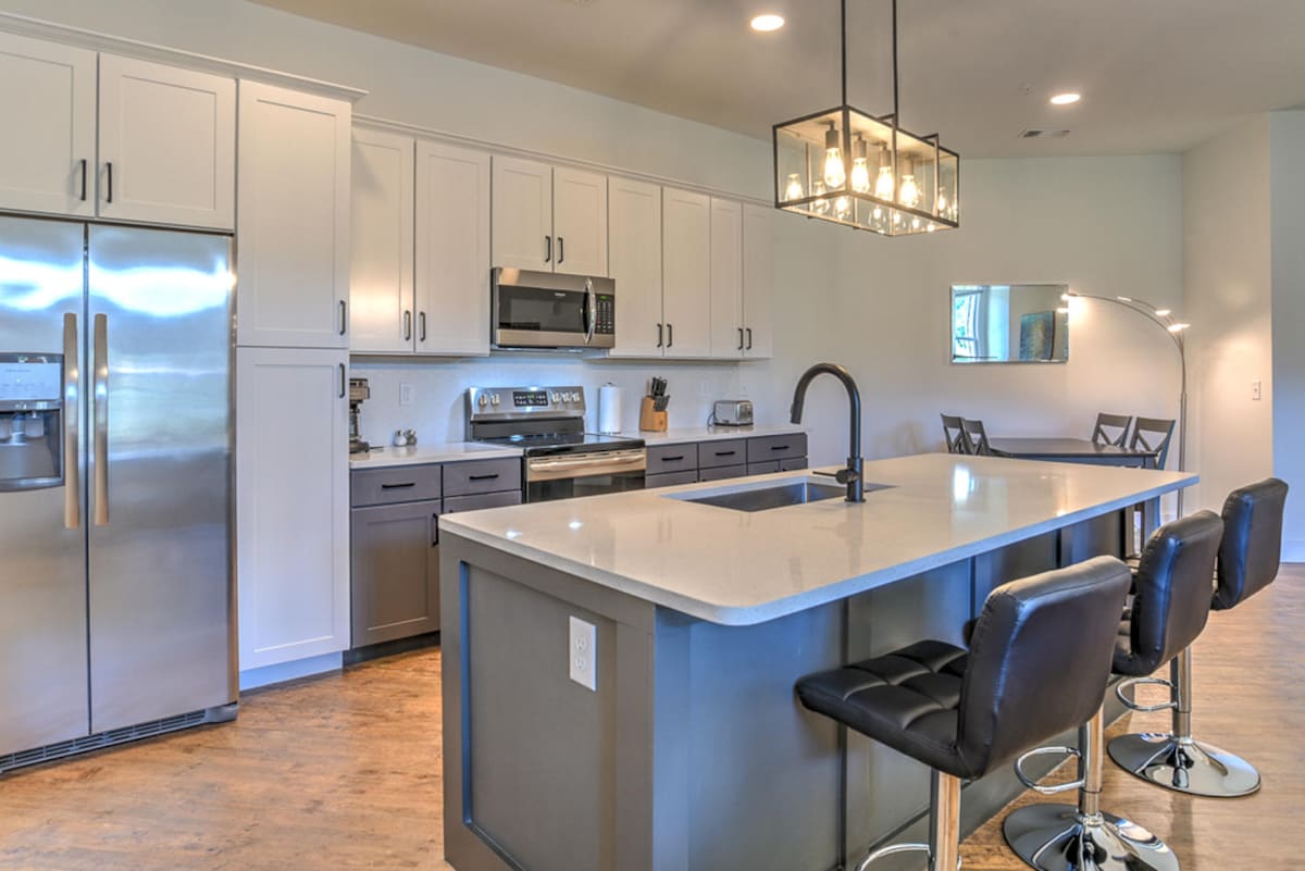 An open kitchen features modern stainless steel appliances, including a refrigerator and microwave. A large island with seating for four is complemented by contemporary fixtures. Ample cabinetry and counter space are visible, along with a dining area in the background, illuminated by natural light.