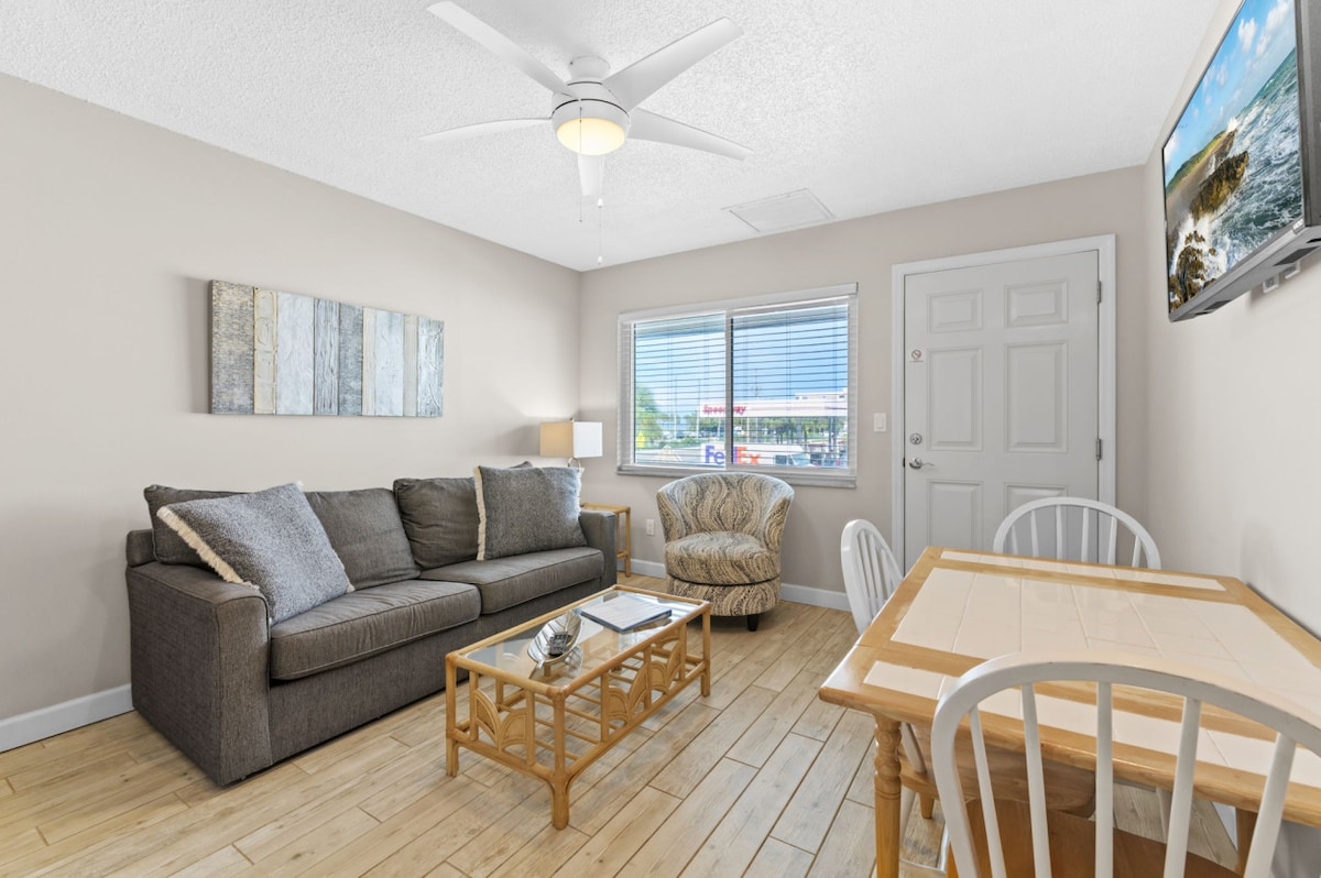 A bright living area features a comfortable gray sofa adorned with decorative pillows, complemented by a wooden coffee table. Nearby, a dining table with four chairs sits under a ceiling fan. Large windows let in natural light, offering a view of the exterior.