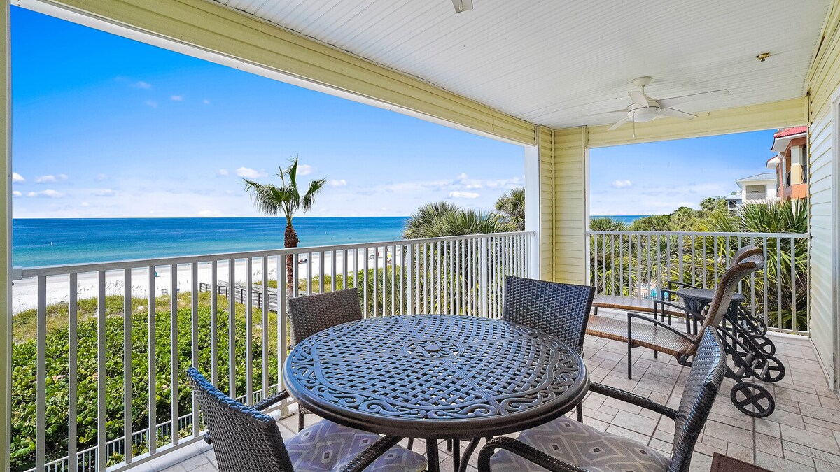 An expansive private balcony features a round dining table surrounded by chairs, offering views of the Gulf of Mexico and palm trees. The outdoor space is shaded by an overhead ceiling fan, providing a relaxing atmosphere for meals or leisure.