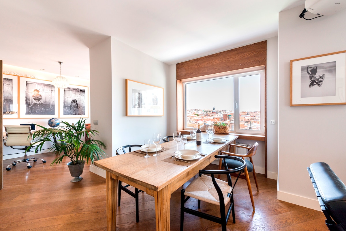 A dining area features a wooden table surrounded by chairs, positioned near a window offering city views. Natural light enters through the window, illuminating the space. Framed artwork adorns the wall, and a potted plant adds a touch of greenery to the setting.