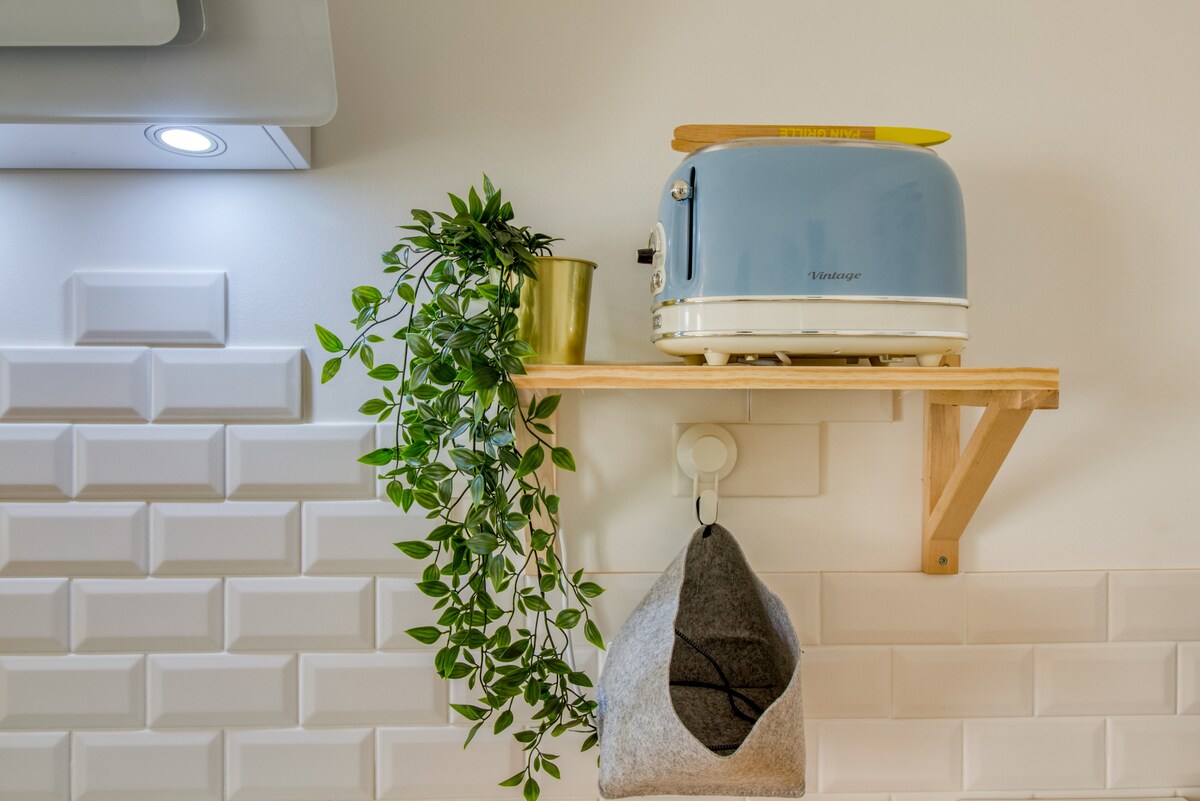 A kitchen shelf displays a mint green toaster alongside a small potted plant. A grey hanging item is positioned below, complementing the textured white tile backdrop. Soft lighting from above enhances the clean lines of the contemporary kitchen design.