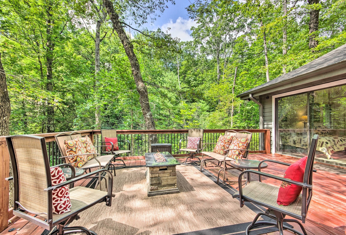 An outdoor deck features comfortable seating arranged around a central fire pit, with soft cushions adorning the chairs. Lush greenery surrounds the space, complemented by a mix of sunlight and shade filtering through the trees, creating a serene retreat.