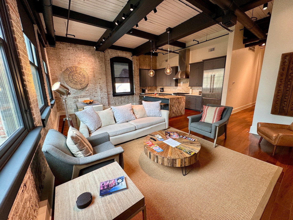 A modern living area features a spacious sofa facing large windows with natural light streaming in. Wooden floors contrast with exposed brick walls. A circular wooden coffee table is placed centrally, surrounded by comfortable seating, and a decorative rug adds warmth to the space.