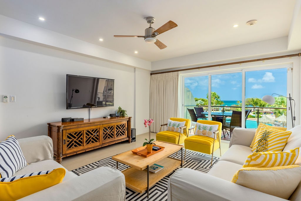 A bright living area features a comfortable arrangement of light-colored sofas and yellow accent chairs. A wooden coffee table sits at the center, with a small plant adding a touch of greenery. Large glass doors open to a balcony, providing beach views and natural light.