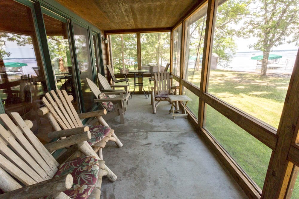 A screened-in porch features several wooden chairs with cushions arranged for relaxation. The floor is concrete, and large windows provide a view of the grassy yard and serene lake in the background. Natural light filters in, enhancing the inviting space.