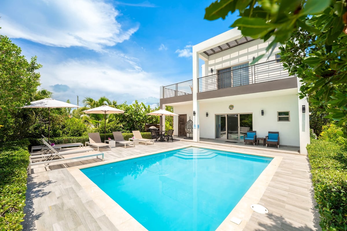 A private pool area is surrounded by lush greenery, featuring sun loungers and large umbrellas for shade. The villa's modern design is highlighted by a spacious outdoor seating area overlooking the pool, complemented by a patio dining set under the cover of the balcony above.