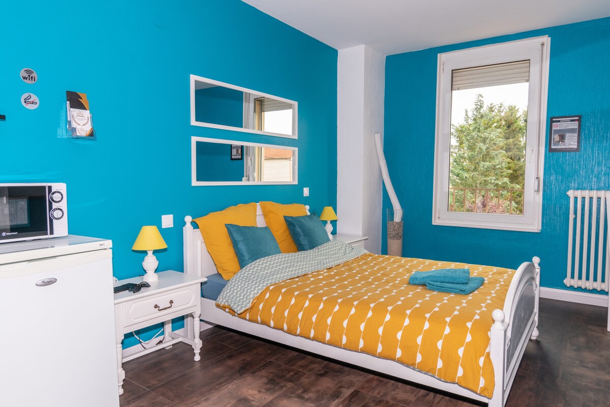 A bright blue wall complements the vibrant yellow bedding arranged on the spacious bed. A mirrored panel reflects natural light from a large window overlooking greenery. The room includes a mini fridge, a bedside table with a lamp, and a cozy seating area.