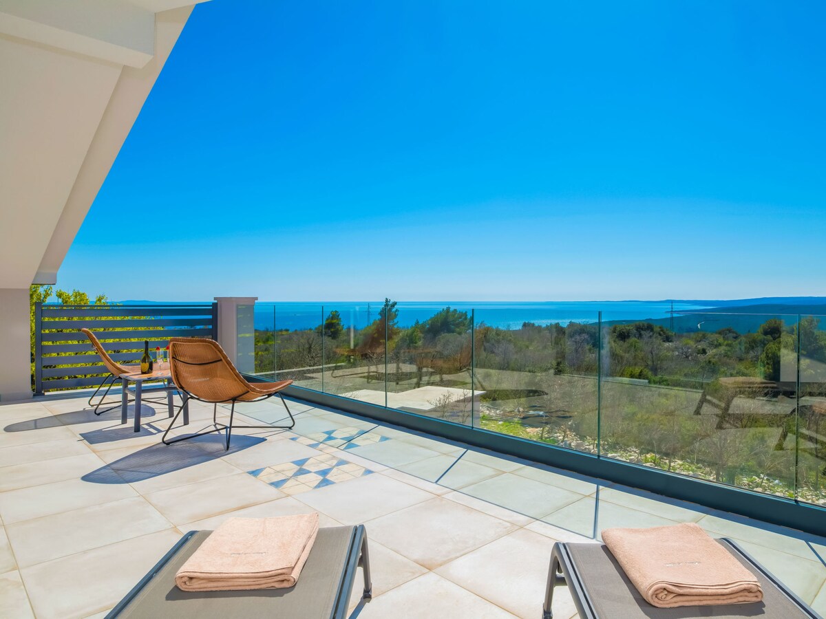A spacious terrace features modern lounge chairs and a small table, offering a clear view of the sea and landscape. Glass railings enhance visibility, allowing the blue sky and distant coastline to be appreciated.