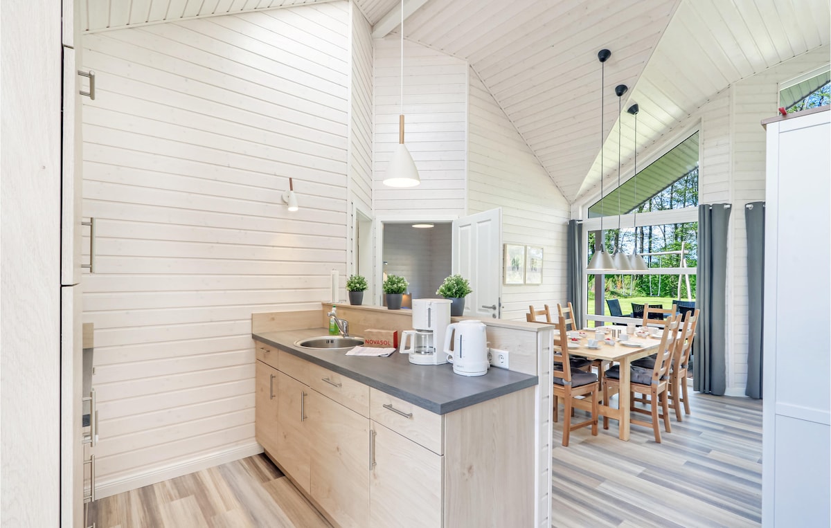 A modern kitchen features light wood cabinetry and a gray countertop. Natural light floods the space through tall windows, illuminating the dining area with a wooden table surrounded by chairs. Potted plants are placed on the counter, adding a touch of greenery.