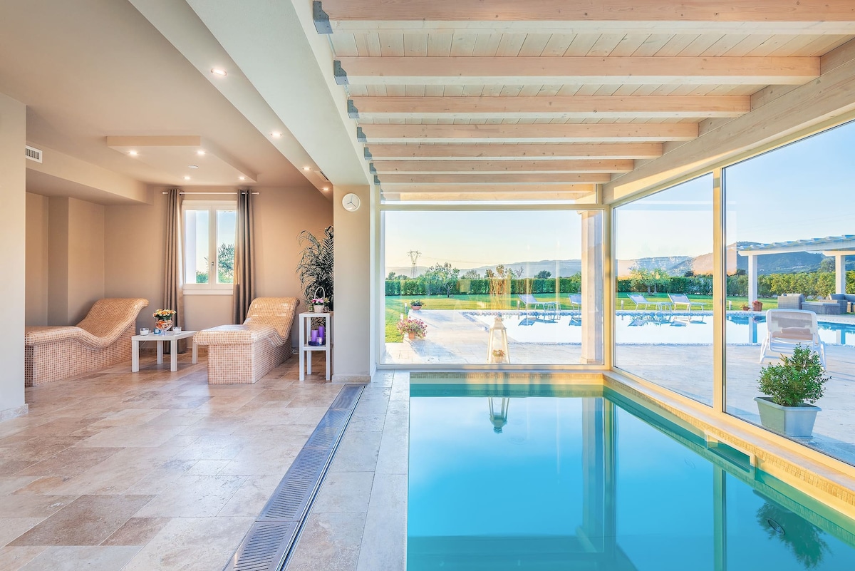 An indoor heated pool is framed by expansive glass walls, offering views of the outdoor garden and pool area. Two modern loungers are positioned nearby, with a small table holding a vase of flowers. Soft lighting enhances the serene ambiance.