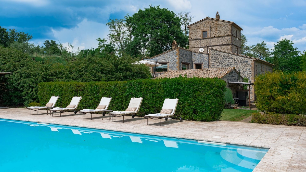 A private pool with a clear blue surface reflects the sky, bordered by a stone patio. Six sun loungers are positioned neatly beside the water. The villa, made of rustic stone, is partially obscured by lush green hedges, offering a sense of seclusion.