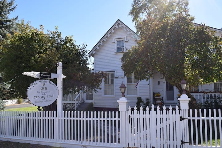 A charming white house features intricate architectural details, nestled among mature trees. A white picket fence surrounds the property, and a welcoming sign indicates the establishment's name. Sunlight filters through the leaves, illuminating the front porch and landscaped yard.