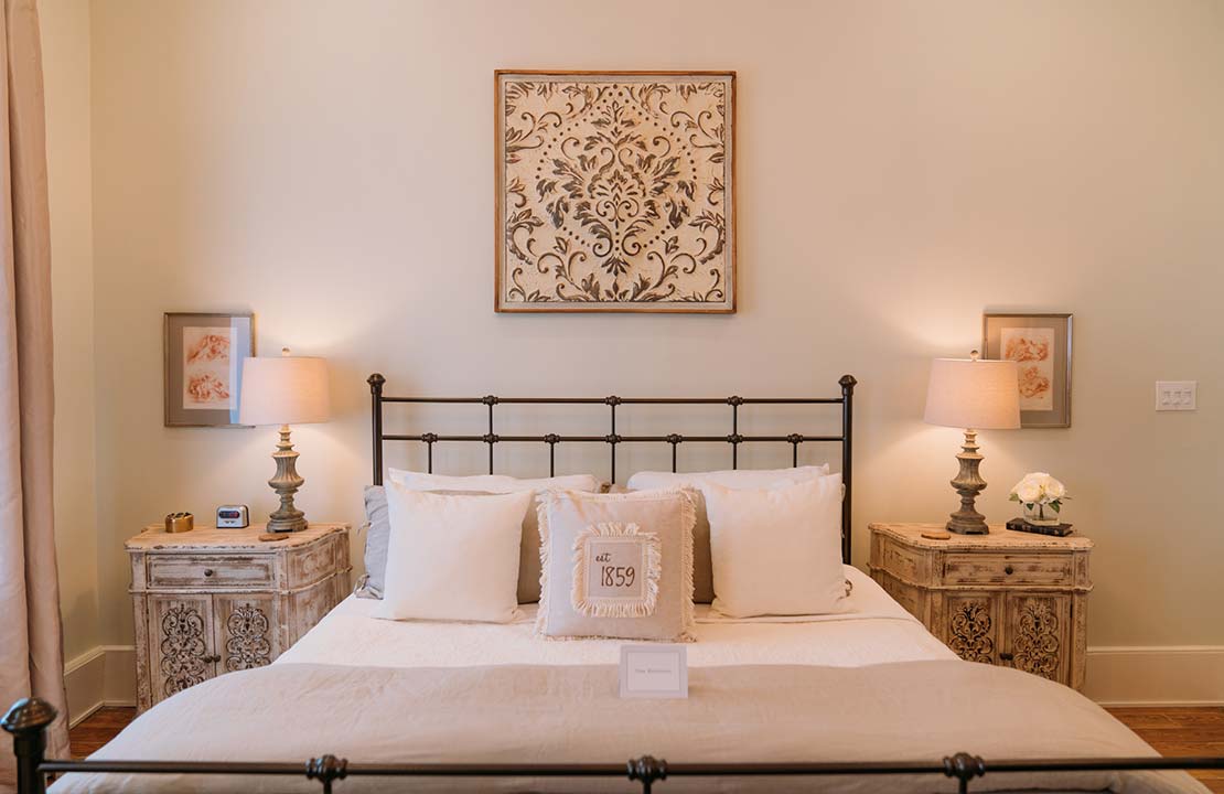 A spacious bed with crisp white linens is centered against a soft-painted wall. Flanking the bed are two wooden bedside tables, each topped with a lamp. Above the bed, a decorative wall art piece adds a touch of elegance. Natural light filters through nearby curtains.