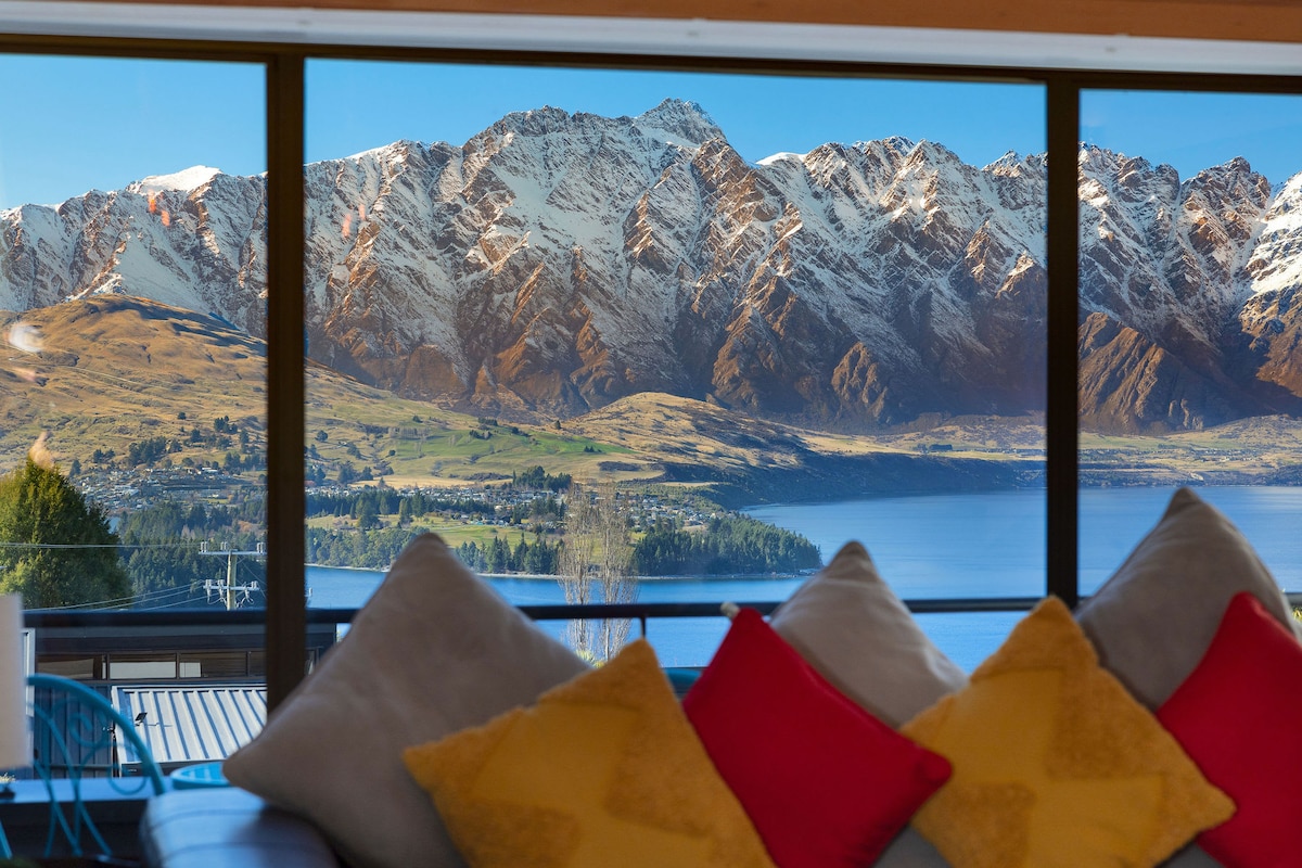Large floor-to-ceiling windows frame stunning views of the Remarkable Mountains and Wakatipu Lake. A cozy seating area features a sofa adorned with bright cushions, inviting relaxation while enjoying the picturesque scenery outside.