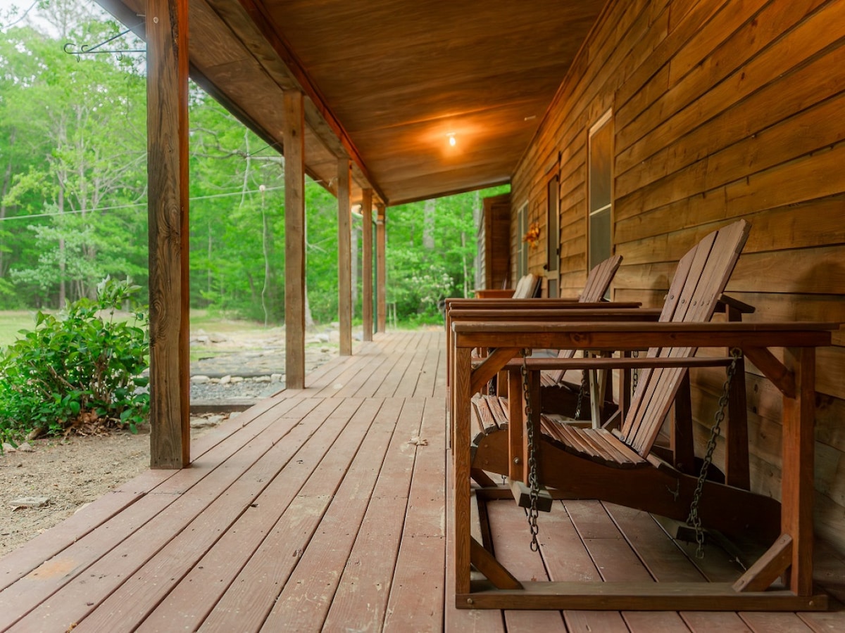 A spacious front porch is lined with rustic wooden chairs, inviting relaxation in nature. The area is surrounded by green foliage, providing a serene backdrop. Warm lighting highlights the wooden beams of the ceiling, creating a welcoming outdoor seating space.