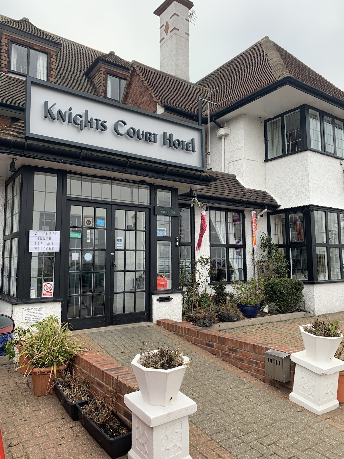 The exterior of the Knights Court Hotel features a charming blend of white and dark brick. Large glass windows allow for natural light, while colorful planters add greenery at the entrance. A sign identifying the property is prominently displayed above the double-door entry.