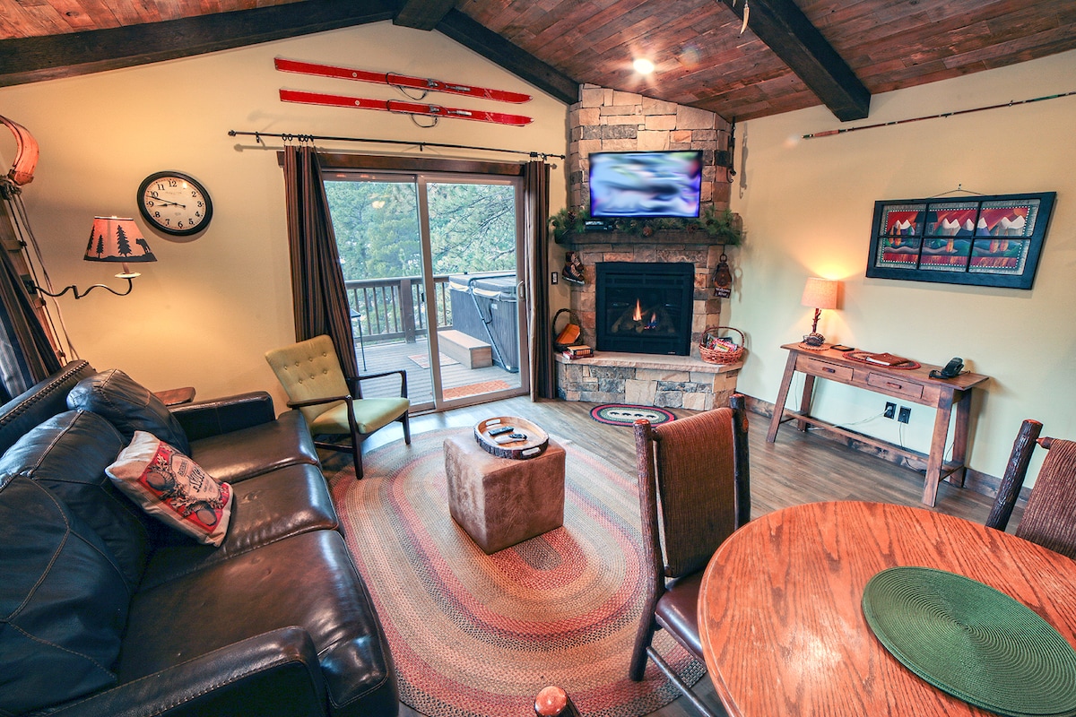 A cozy living room is furnished with a dark leather sofa and a green armchair. A stone fireplace is set against a wooden wall, with a flat-screen TV mounted above. A round dining table sits nearby, illuminated by natural light from a sliding door that opens to a deck.