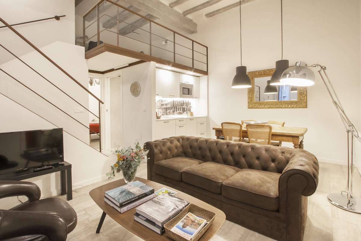 A cozy living area features a tufted brown sofa, a wooden coffee table with magazines, and a TV. A dining table with chairs sits nearby, and a kitchenette is visible in the background. Soft lighting is provided by stylish lamps and a decorative mirror.