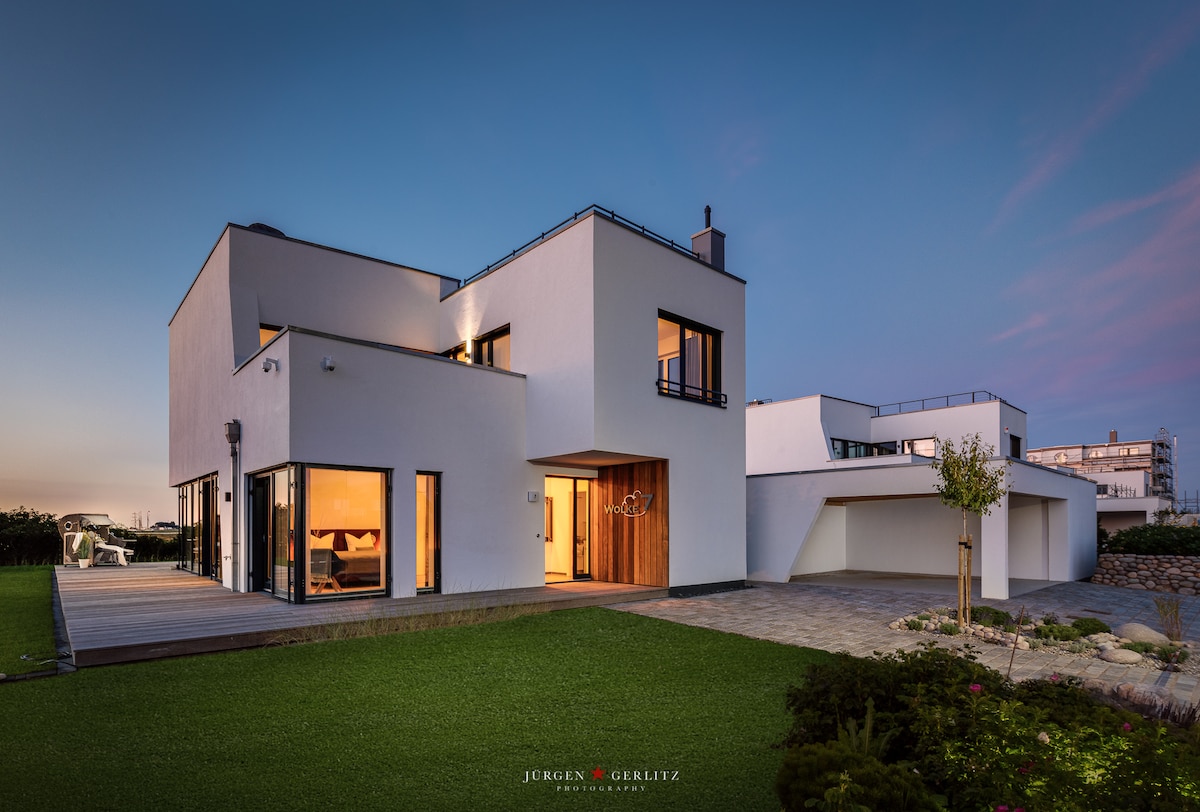 A modern two-story house is showcased with clean lines and large windows. The structure features an expansive wooden deck leading to a well-maintained green lawn. Adjacent buildings are visible in the background, and soft evening light adds a serene tone to the scene.