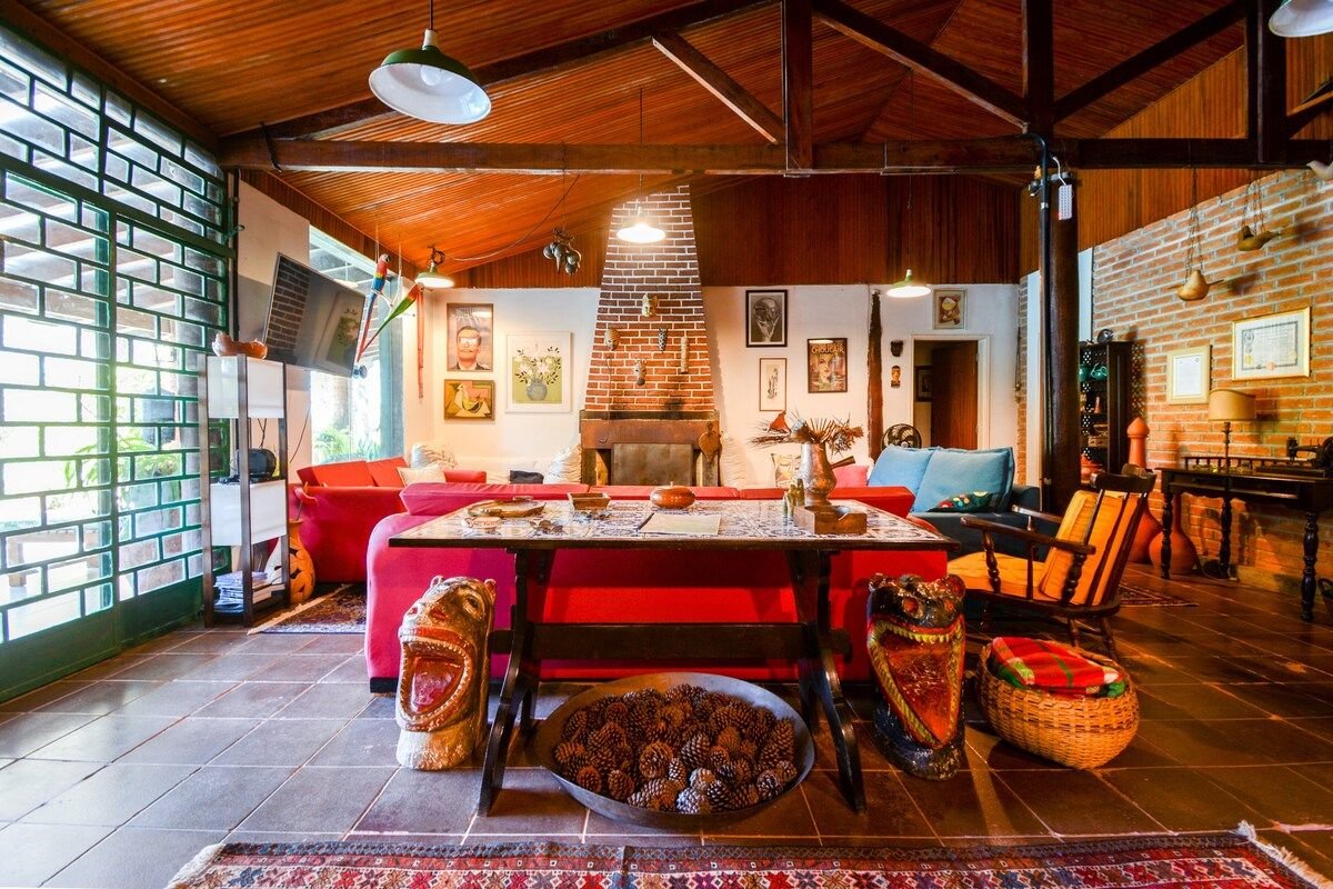 An inviting living area showcases high wooden ceilings and large windows, allowing natural light to fill the space. A central table is surrounded by vibrant seating, with decorative accents including artistic sculptures and a cozy fireplace, enhancing the room's ambiance.