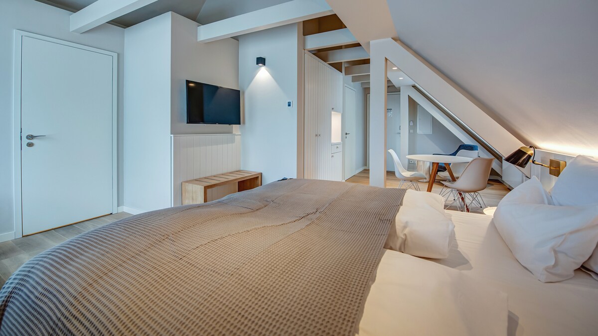 A spacious hotel room showcases a large bed adorned with a textured blanket and plush pillows. A flat-screen television is mounted on the wall, and a contemporary dining area is visible nearby, illuminated by natural light from the window.