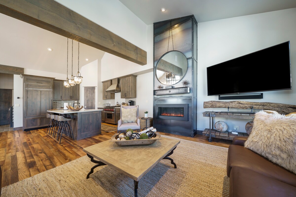 The living area features high ceilings with exposed beams and a large circular mirror above an elegant gas fireplace. A dark leather sofa and wooden coffee table complement the hardwood flooring, while a generously sized kitchen is visible in the background.